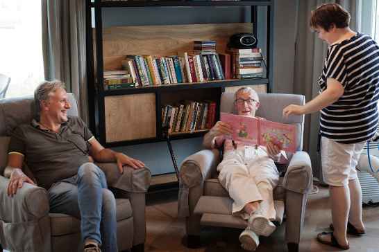 Drie mensen zitten en staan in een woonkamer met stoelen en een boekenkast. Eén persoon bekijkt een boek.