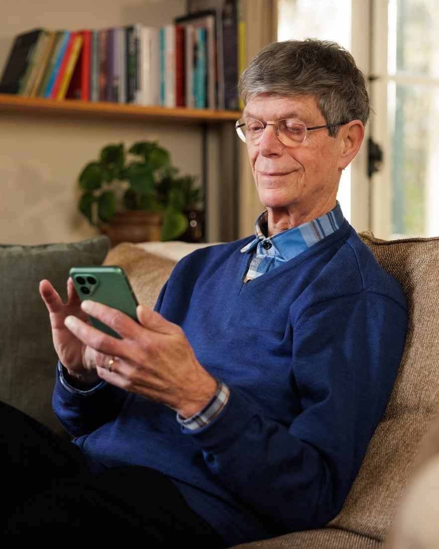 Een man zit op een bank en kijkt op zijn telefoon in een woonkamer met boeken en planten.