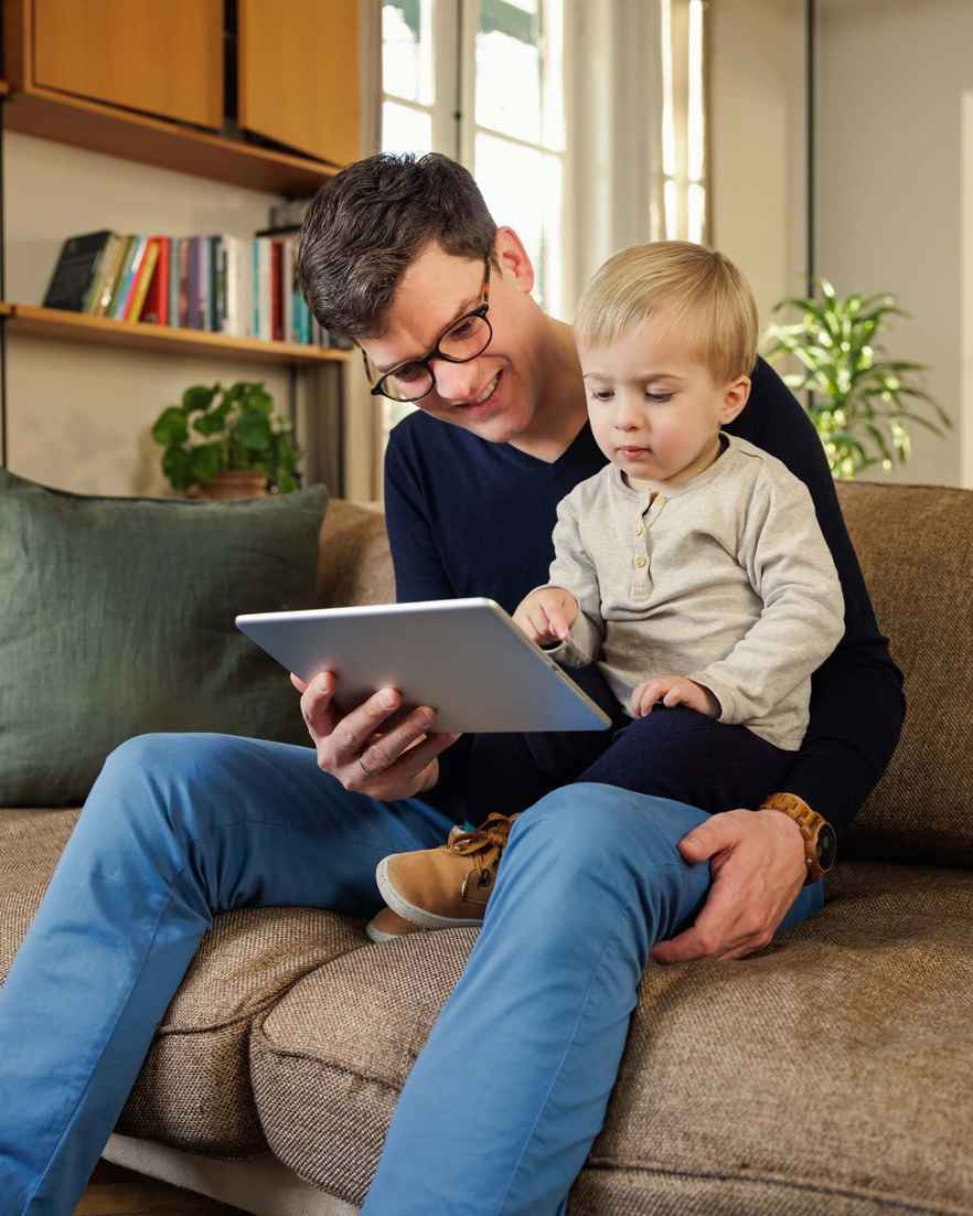 Een man met een bril en een kind op schoot zitten samen op een bank en kijken naar een tablet.