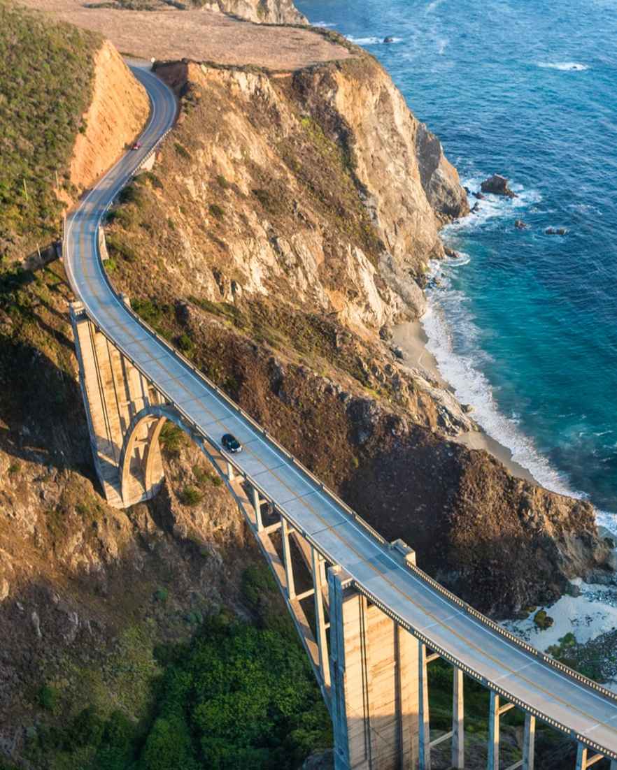Een auto rijdt over een lange brug langs de rotsen boven de zee. De weg volgt de kust en buigt mee met het landschap.