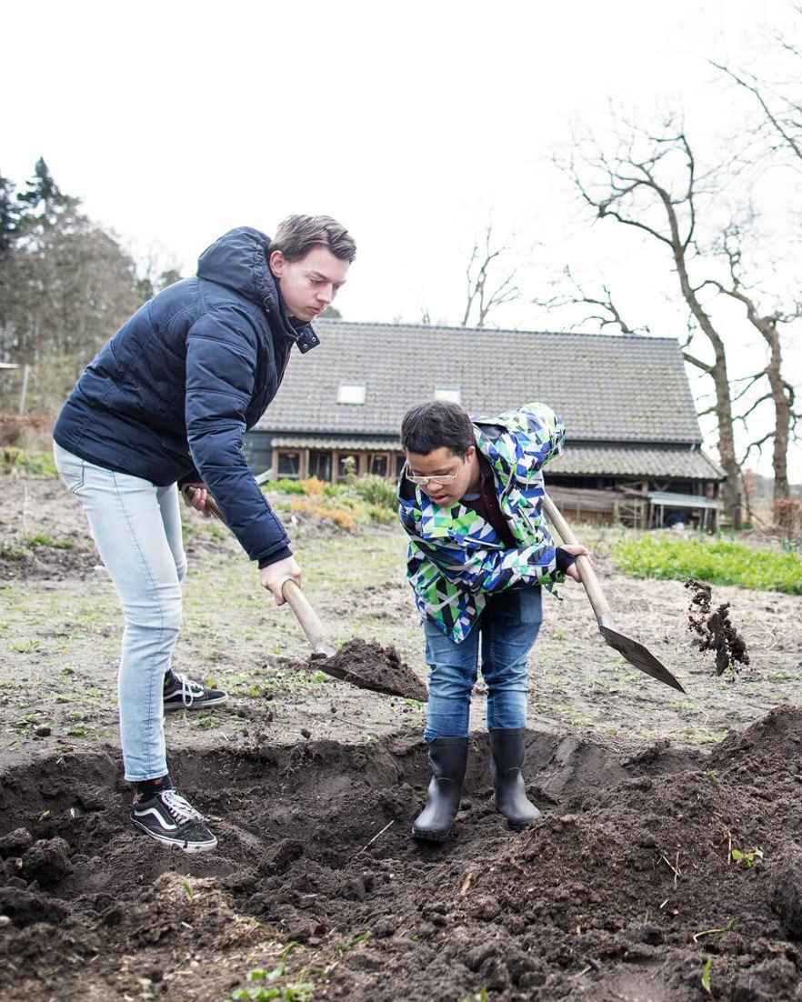 Een volwassene en een kind graven samen in de grond met een schep. Ze werken buiten op een open stuk land bij een gebouw.