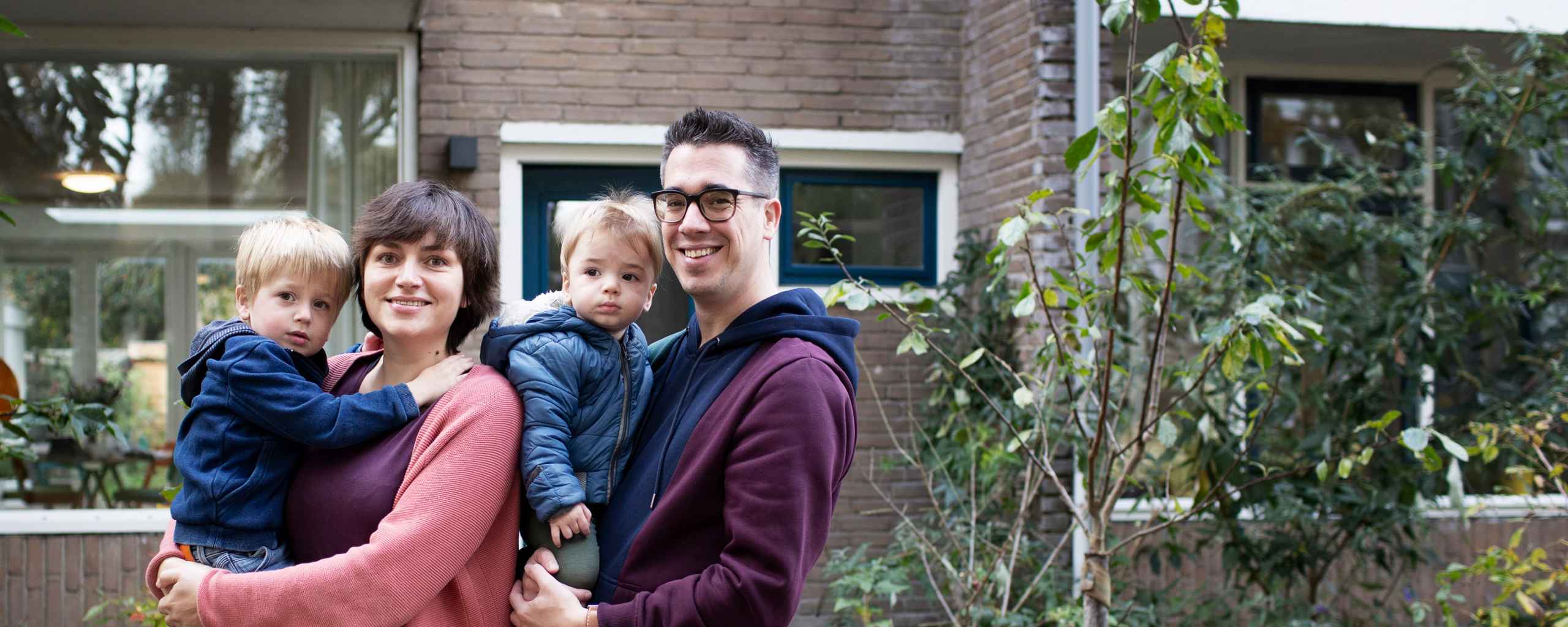 Een gezin lacht voor een bakstenen huis. De vader houdt een kind in een blauw jack vast. Hij draagt een paarse trui. De moeder houdt ook een kind vast. Achter hen groeien planten en bomen.