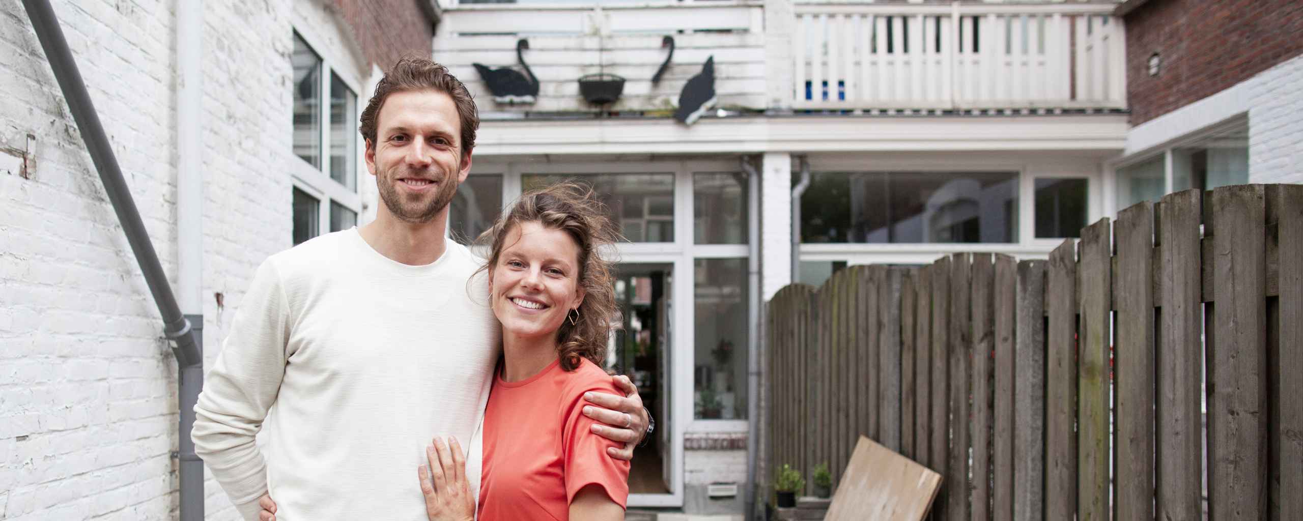 Een man en een vrouw lachen voor een wit huis met een houten schutting. De man draagt een licht shirt en de vrouw een roze shirt. Achter hen hangen bloempotten aan de muur.