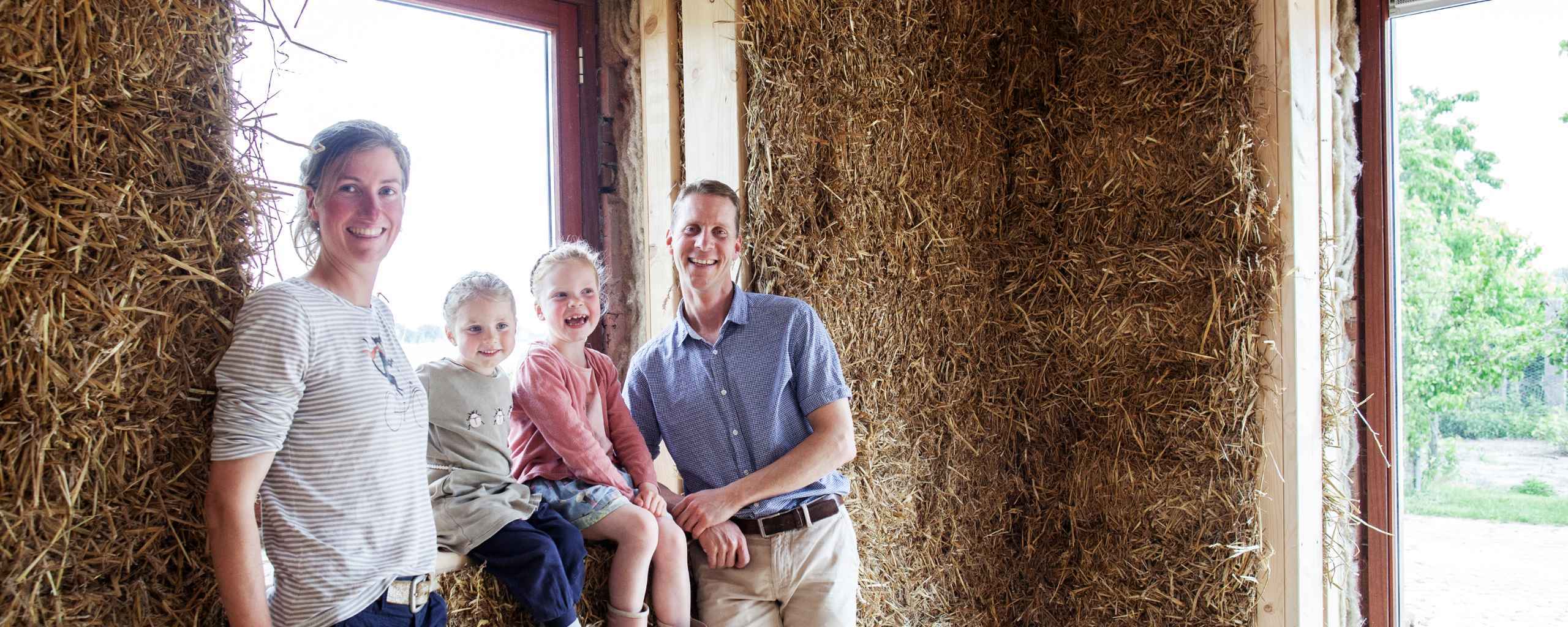 Een vrouw en een man lachen met twee kinderen in een schuur. Achter hen ligt een grote berg stro. De kinderen zitten op een houten bank en kijken blij.