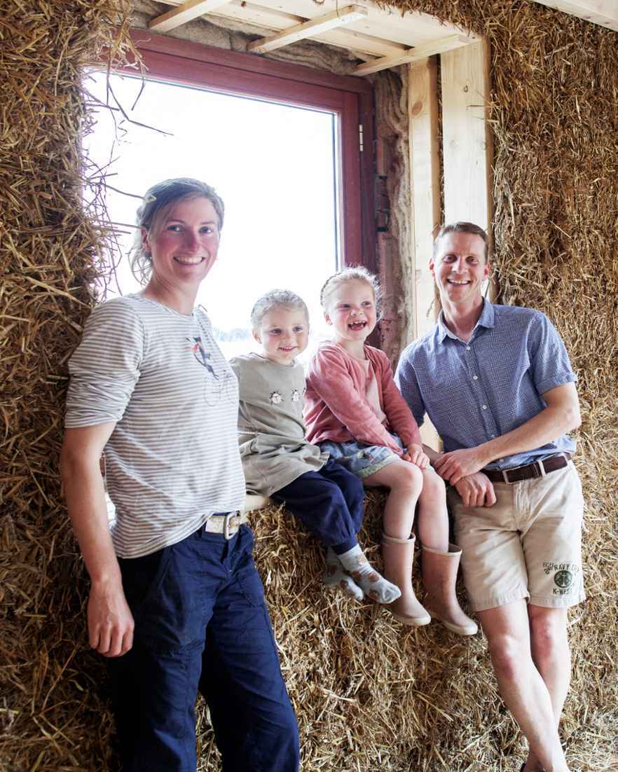 Een vrouw en een man lachen met twee kinderen in een schuur. Achter hen ligt een grote berg stro. De kinderen zitten op een houten bank en kijken blij.