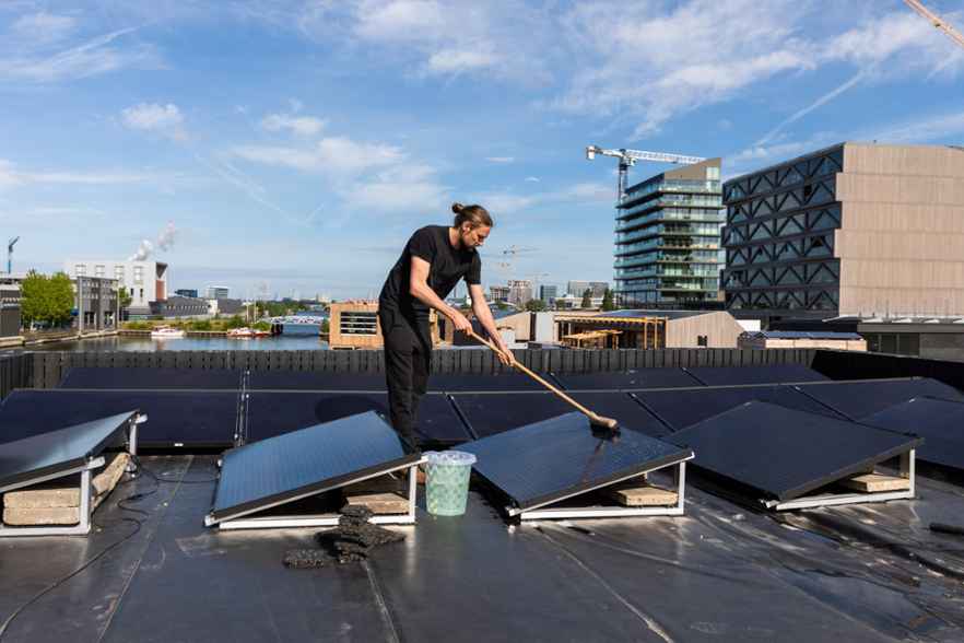 Een man maakt zonnepanelen schoon op een dak. Achter hem liggen water, boten, moderne gebouwen en hijskranen.