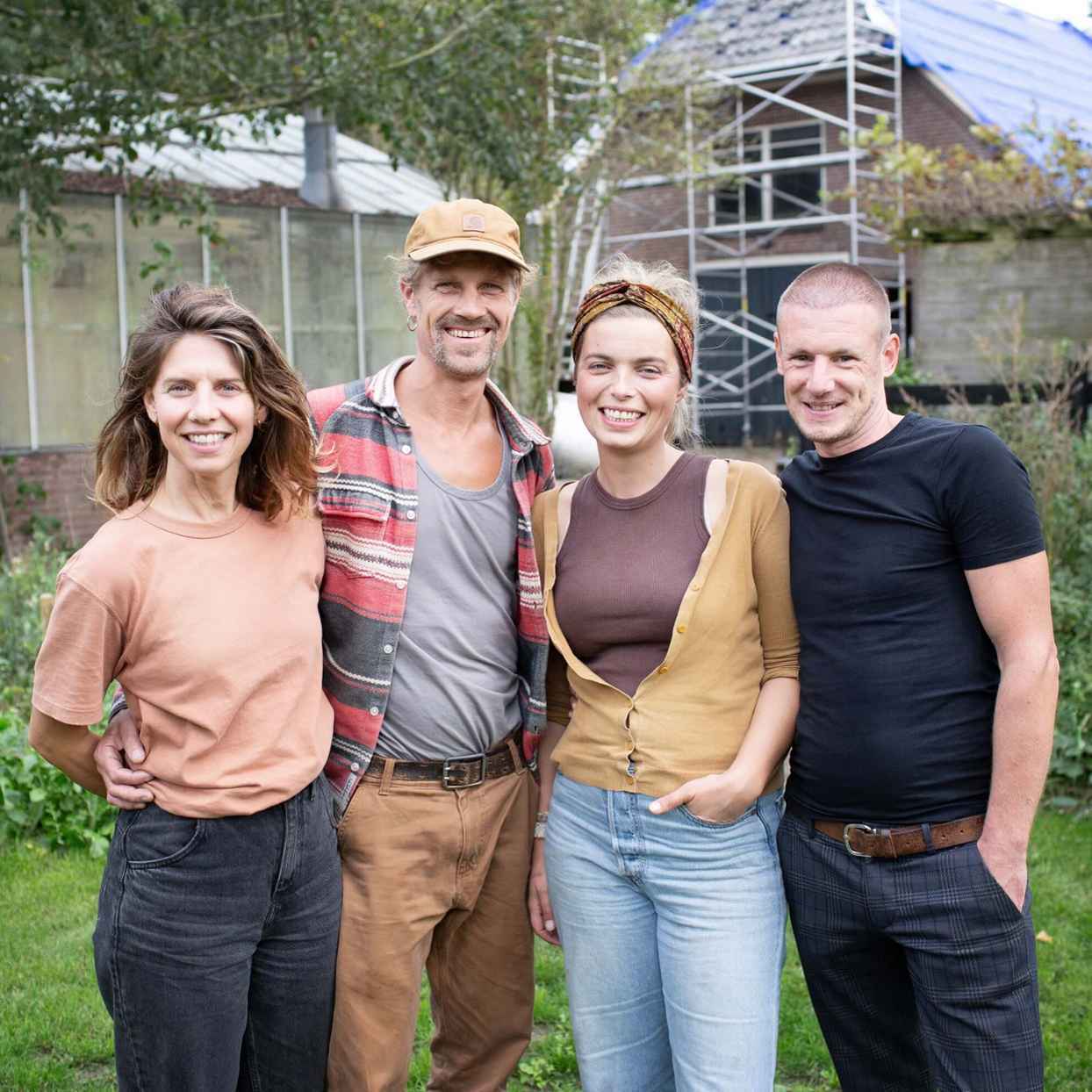 Vier mensen staan samen in een tuin. Ze hebben hun armen om elkaar heen en kijken naar de camera. Op de achtergrond staan een kas, bomen en een huis met steigers.
