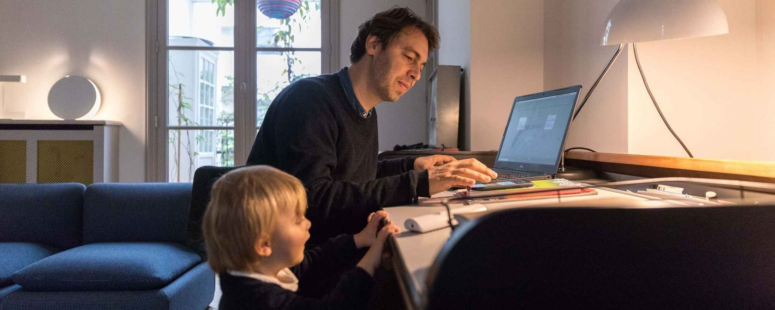 Een man werkt op een laptop aan een bureau. Een kind staat naast hem in de kamer.