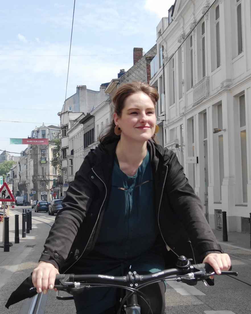 Een persoon fietst door een smalle straat in een stad. Langs de straat staan winkels, huizen en paaltjes.