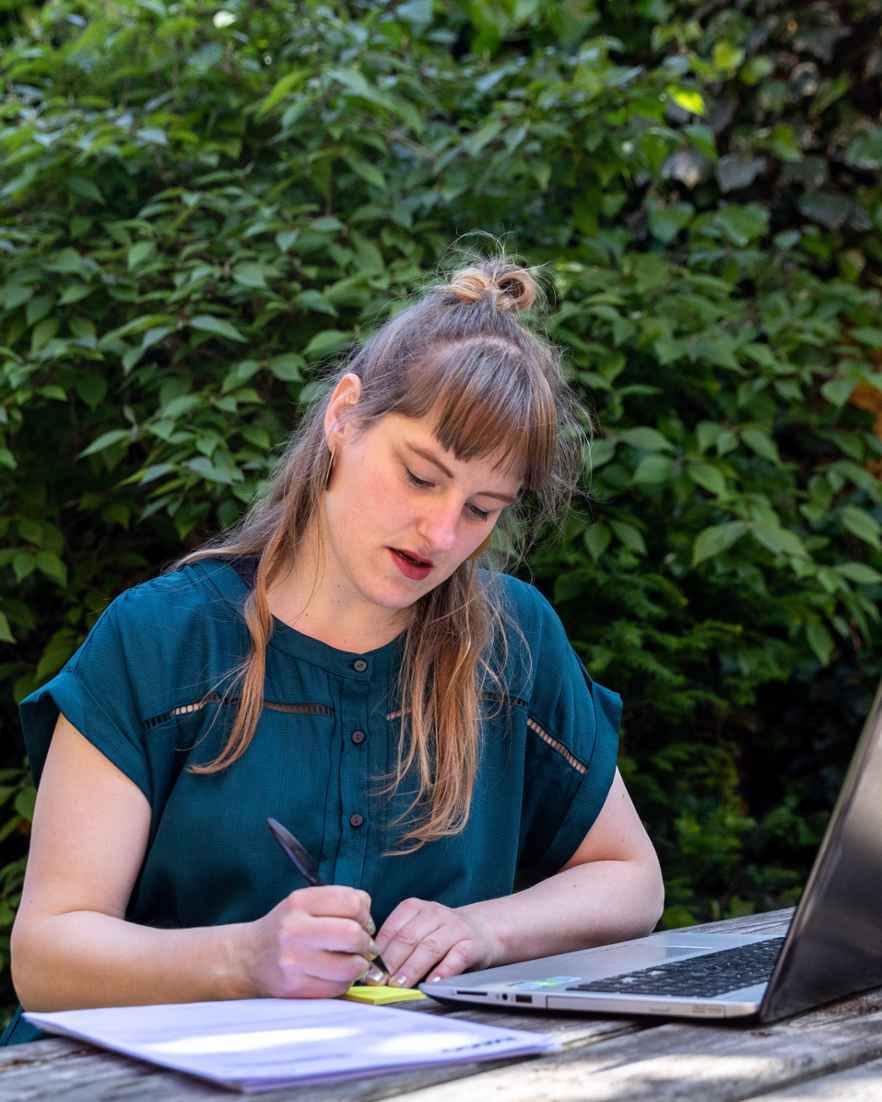 Iemand zit aan een houten tafel in een groene tuin en schrijft in een notitieboek. Op tafel staat ook een open laptop en een klein plantje.