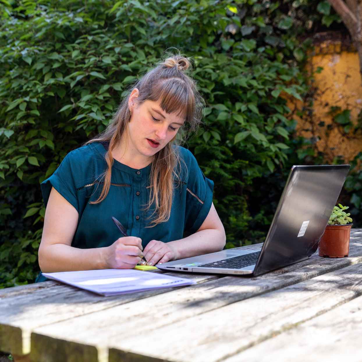 Iemand zit aan een houten tafel in een groene tuin en schrijft in een notitieboek. Op tafel staat ook een open laptop en een klein plantje.