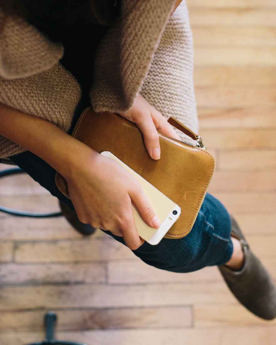 Bovenaanzicht van iemand in spijkerbroek en enkellaarsjes, zittend op een krukje met een telefoon en een leren hoes op schoot.