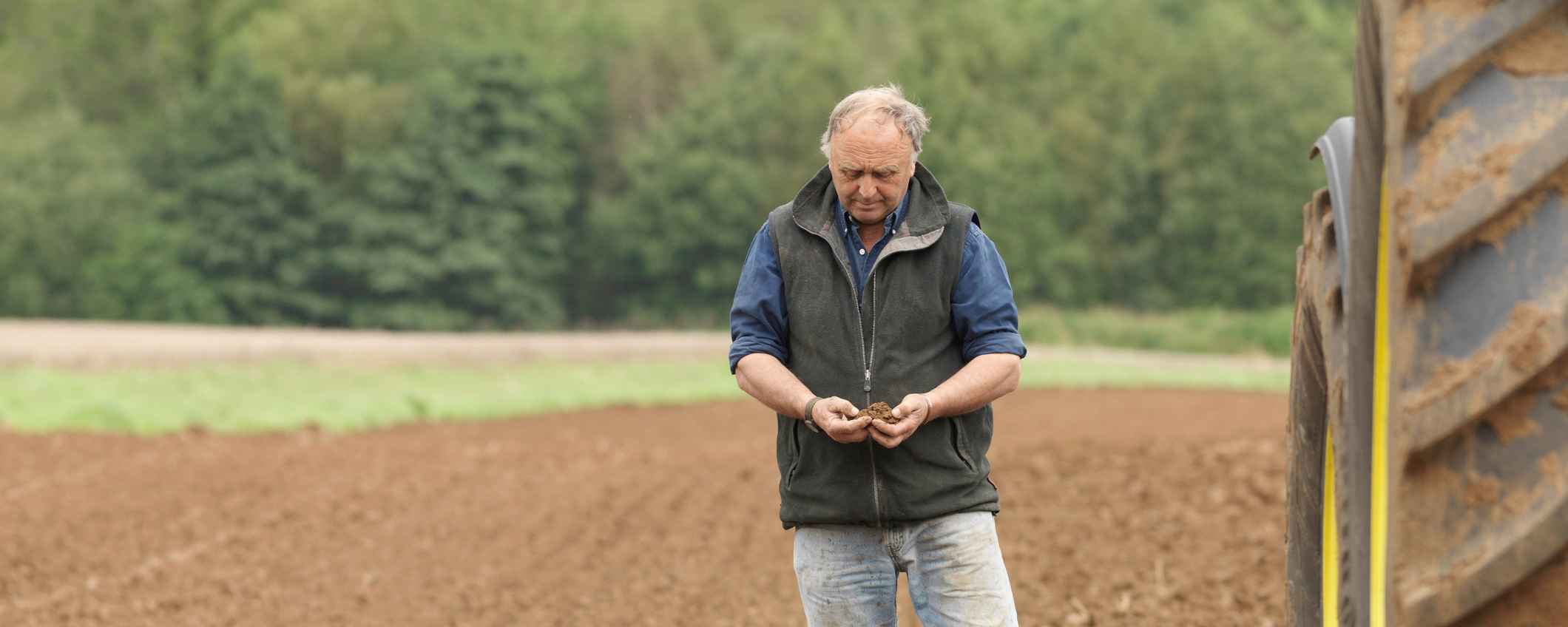 Een boer staat op een akker en houdt aarde in zijn handen. Naast hem staat een grote tractor.