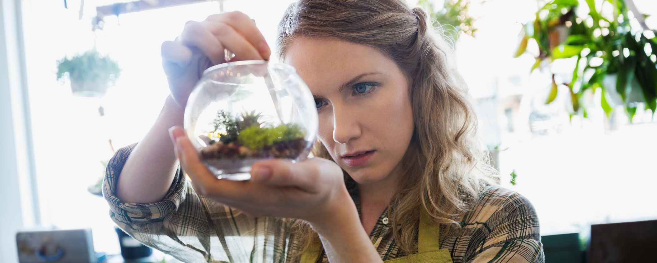 Iemand houdt een glazen potje met kleine planten vast en bekijkt het aandachtig. Op de achtergrond hangen meer planten.