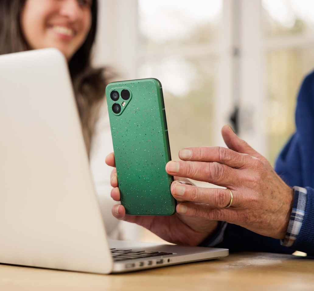 Man houdt een groene telefoon vast naast een open laptop aan een tafel. Naast de man zit een vrouw.