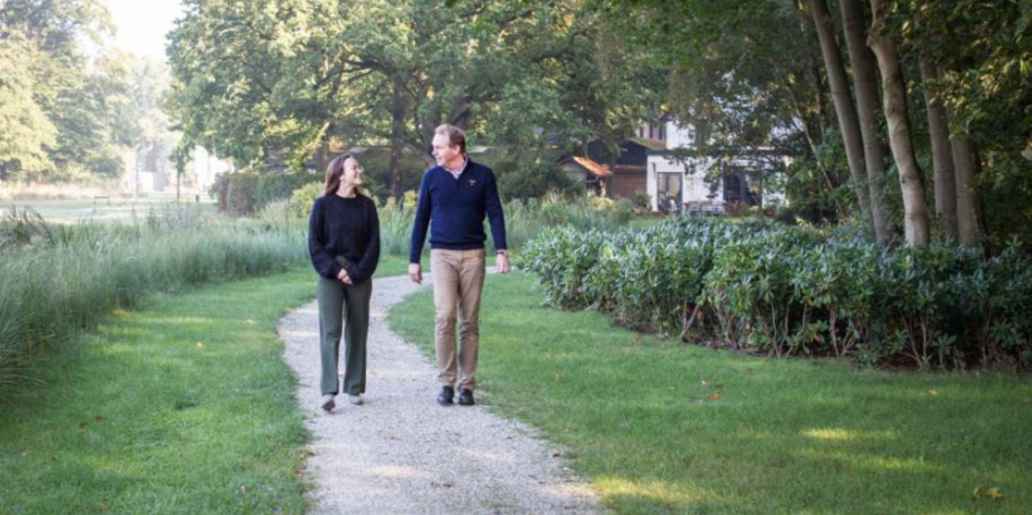 Twee mensen wandelen samen over een pad in een park. Langs het pas staan gras, struiken en bomen.