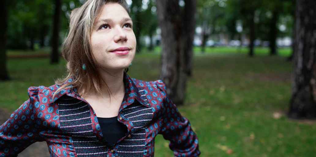 Een vrouw staat in een park met bomen en gras. De vrouw draagt een kleurrijke blouse met een patroon.