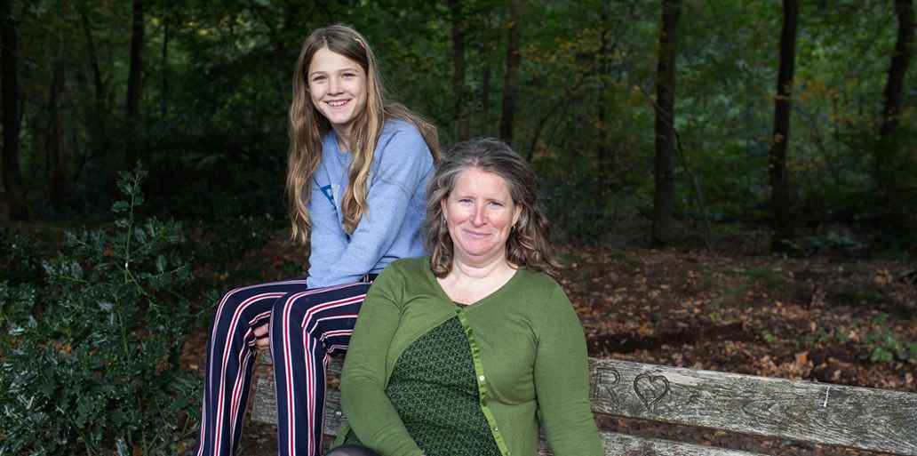 Twee mensen zitten samen op een houten bank in een bos. Rondom hen staan bomen en bladeren op de grond.
