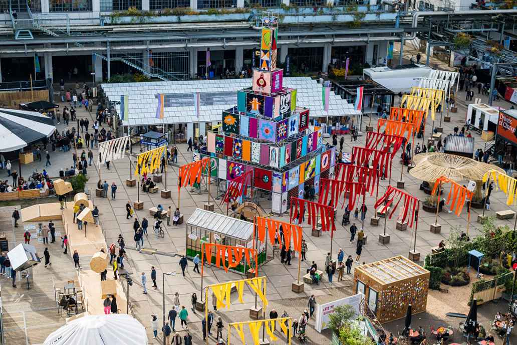 Mensen lopen over een groot plein tijdens Dutch Design Week, met een kleurrijke kunstinstallatie en opvallende banen van stof.