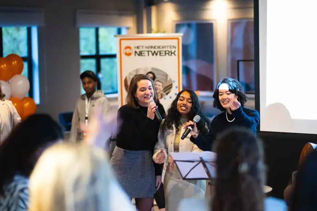 Drie vrouwen staan op een podium met microfoons tijdens een bijkomst, terwijl publiek toekijkt.