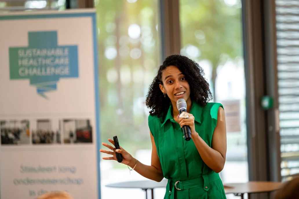 Een vrouw geeft een presentatie en spreekt in een microfoon naast een banner over duurzame zorg.