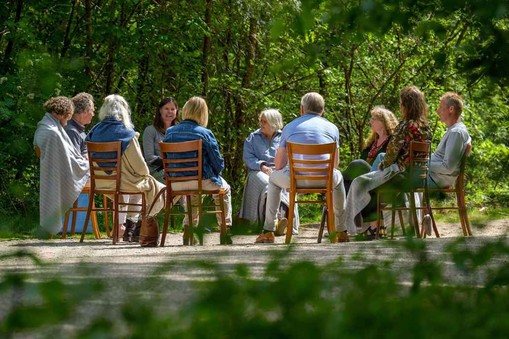Een groep mensen zit buiten in een kring op stoelen, midden in een groene bosrijke omgeving.