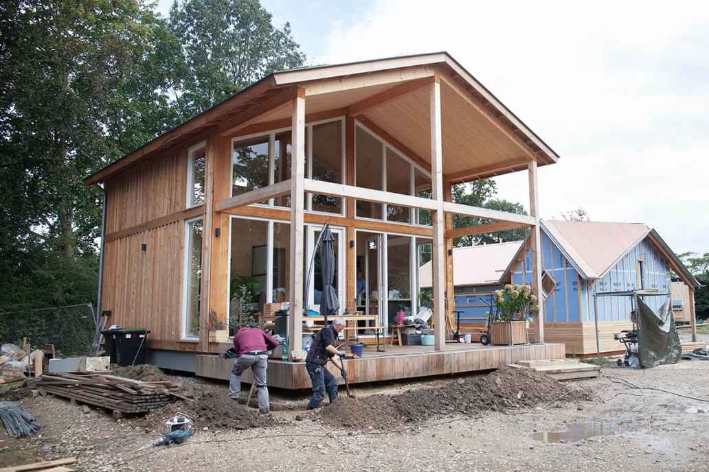 Twee mensen werken buiten aan een houten huis met grote ramen een veranda. Op de achtergrond staat nog een huis in aanbouw.
