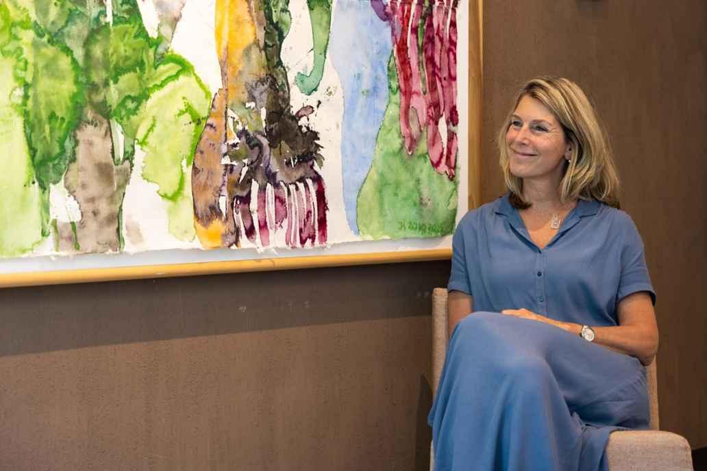 Riëlla Hollander, managing director TRMC. Riëlla draagt een lichtblauwe bloesjurk en zit op een stoel. Ze heeft halflang blond haar en glimlacht. Achter haar hangt een abstract aquarel schilderij aan een bruine leemmuur.