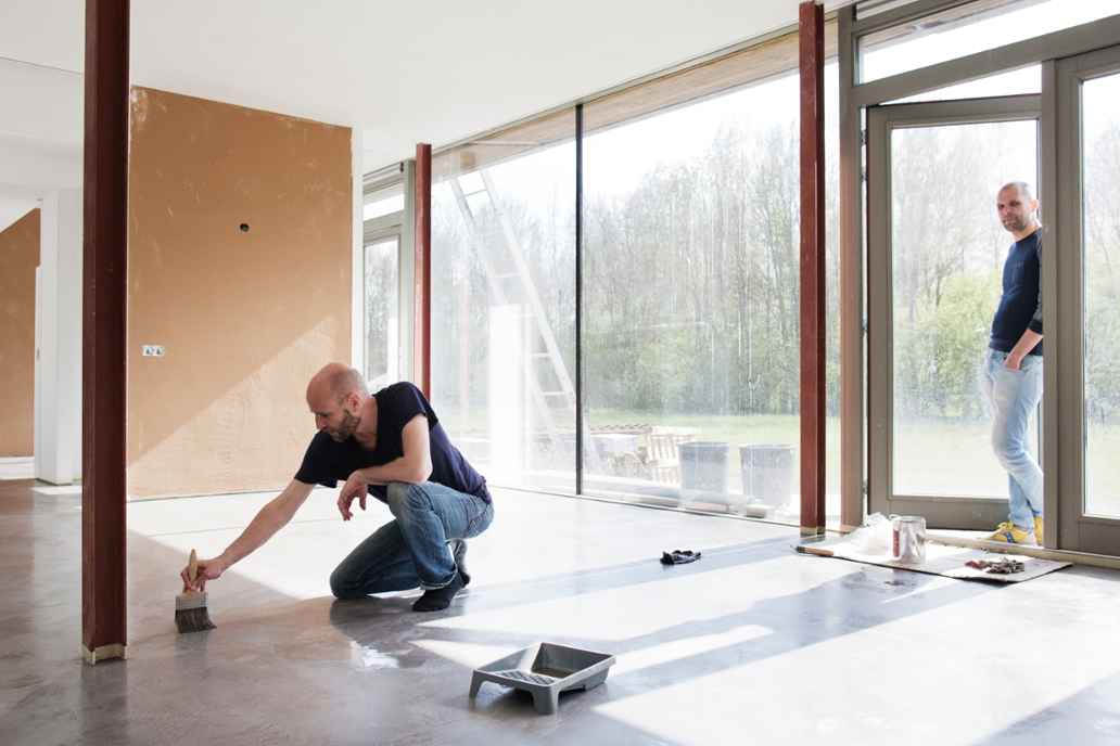 Een man schildert de vloer in een lichte ruimte. Een andere man kijkt toe bij de grote glazen deuren.
