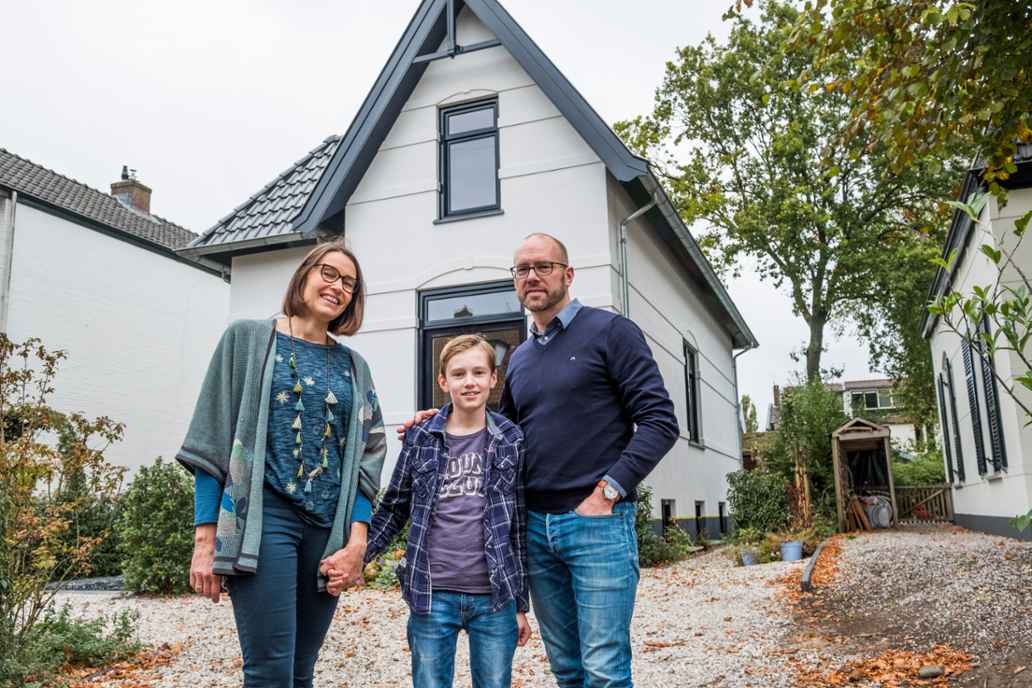 Drie mensen staan voor een wit huis met een tuin. Een kind staat tussen twee volwassenen en houdt een hand vast.
