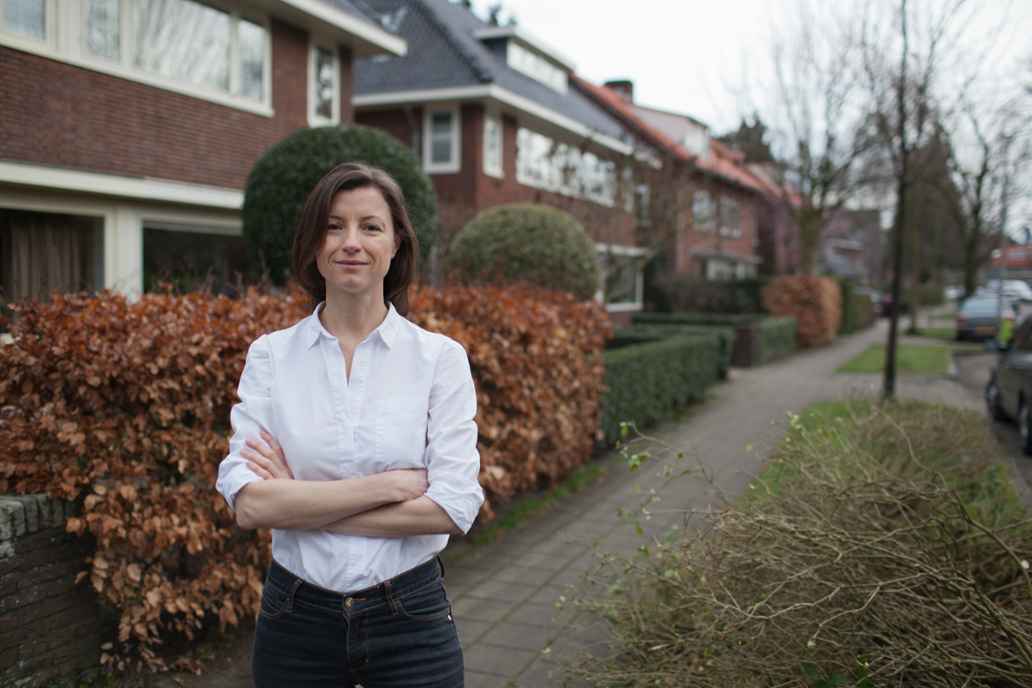 Een vrouw staat met haar armen over elkaar glimlachend in een straat met huizen, hagen en bomen.