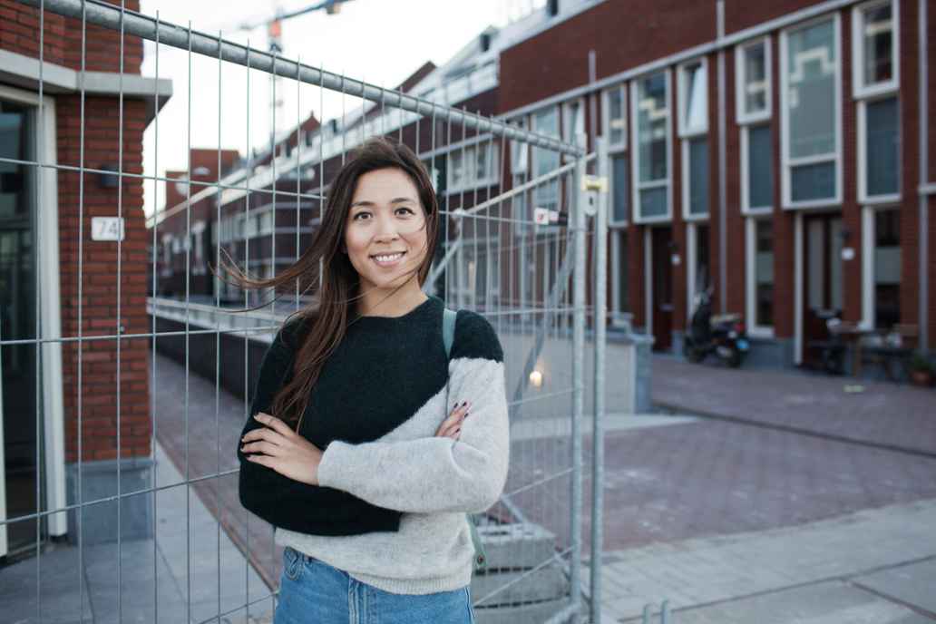 Een vrouw staat voor een hek in een straat met huizen. De wind waait door haar haar.