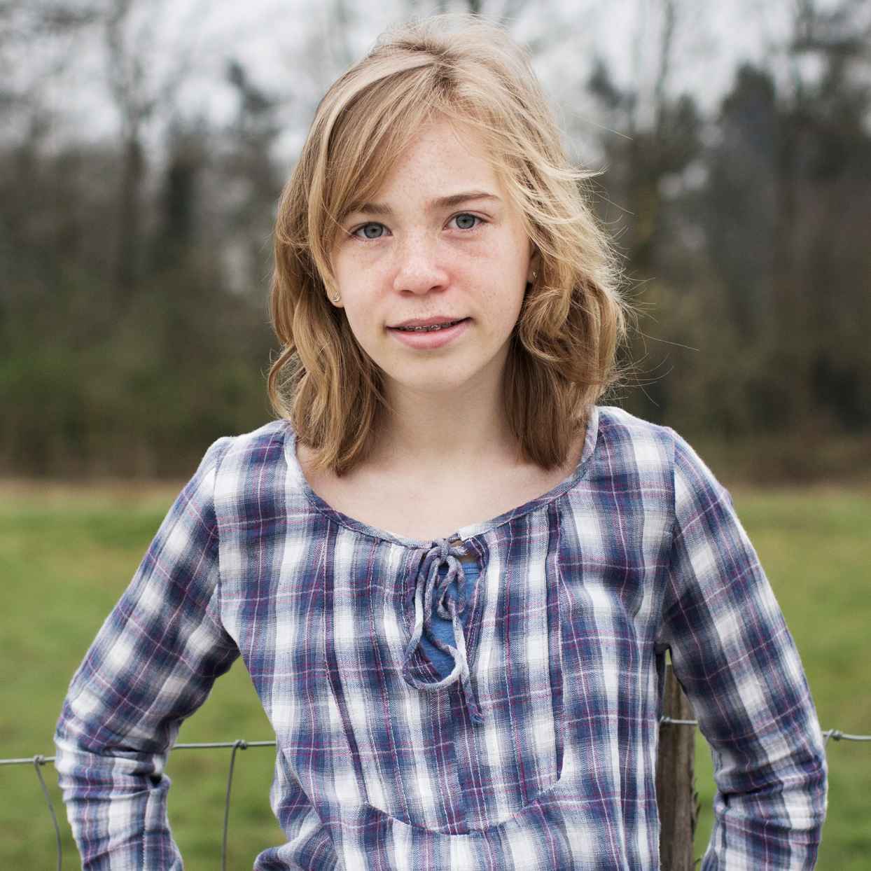 Een tienermeisje staat buiten op een grasveld en draagt een blauw‑geruite blouse.