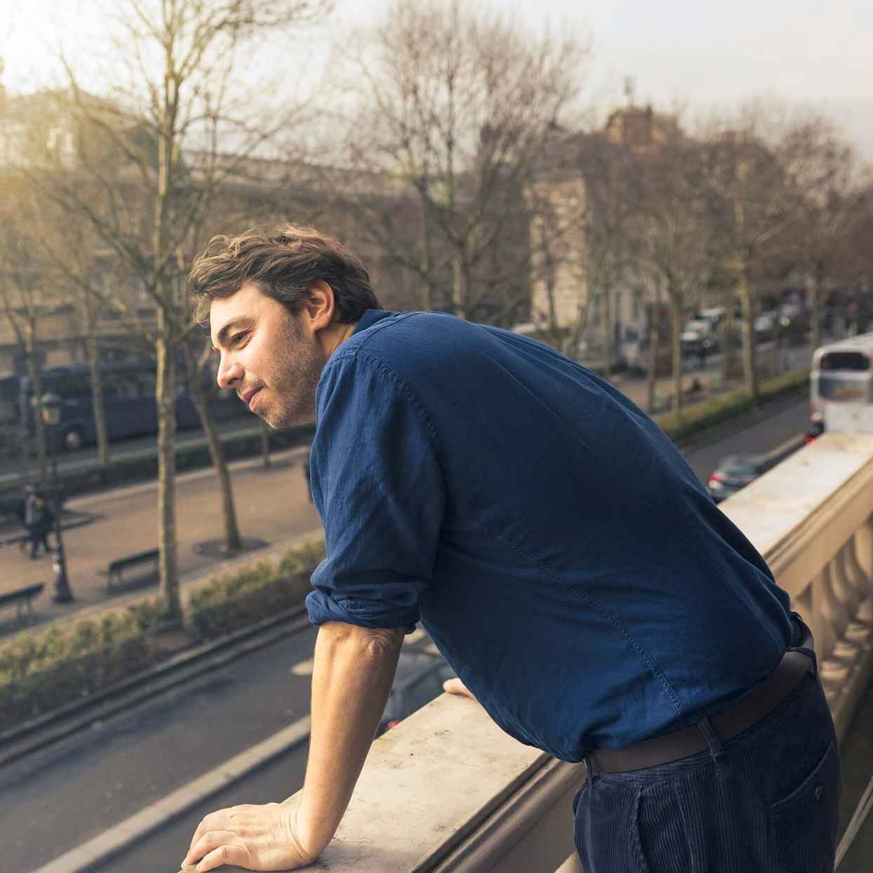 Een man leunt over een balkon en kijkt uit over een straat met bomen.