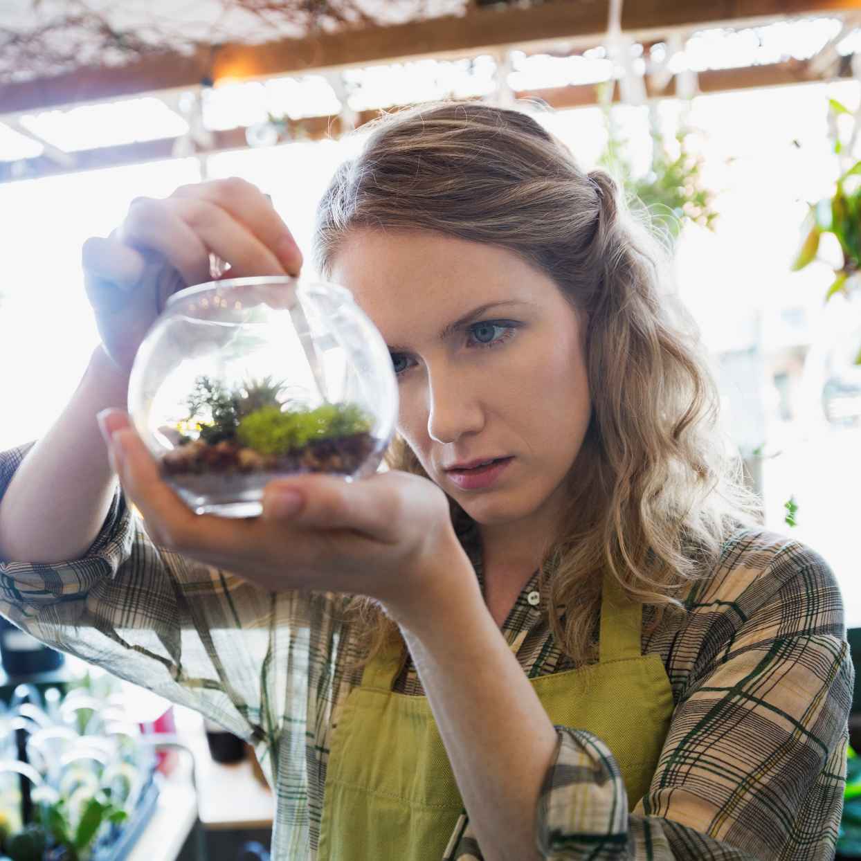 Iemand houdt een glazen potje met kleine planten vast en bekijkt het aandachtig. Op de achtergrond hangen meer planten.