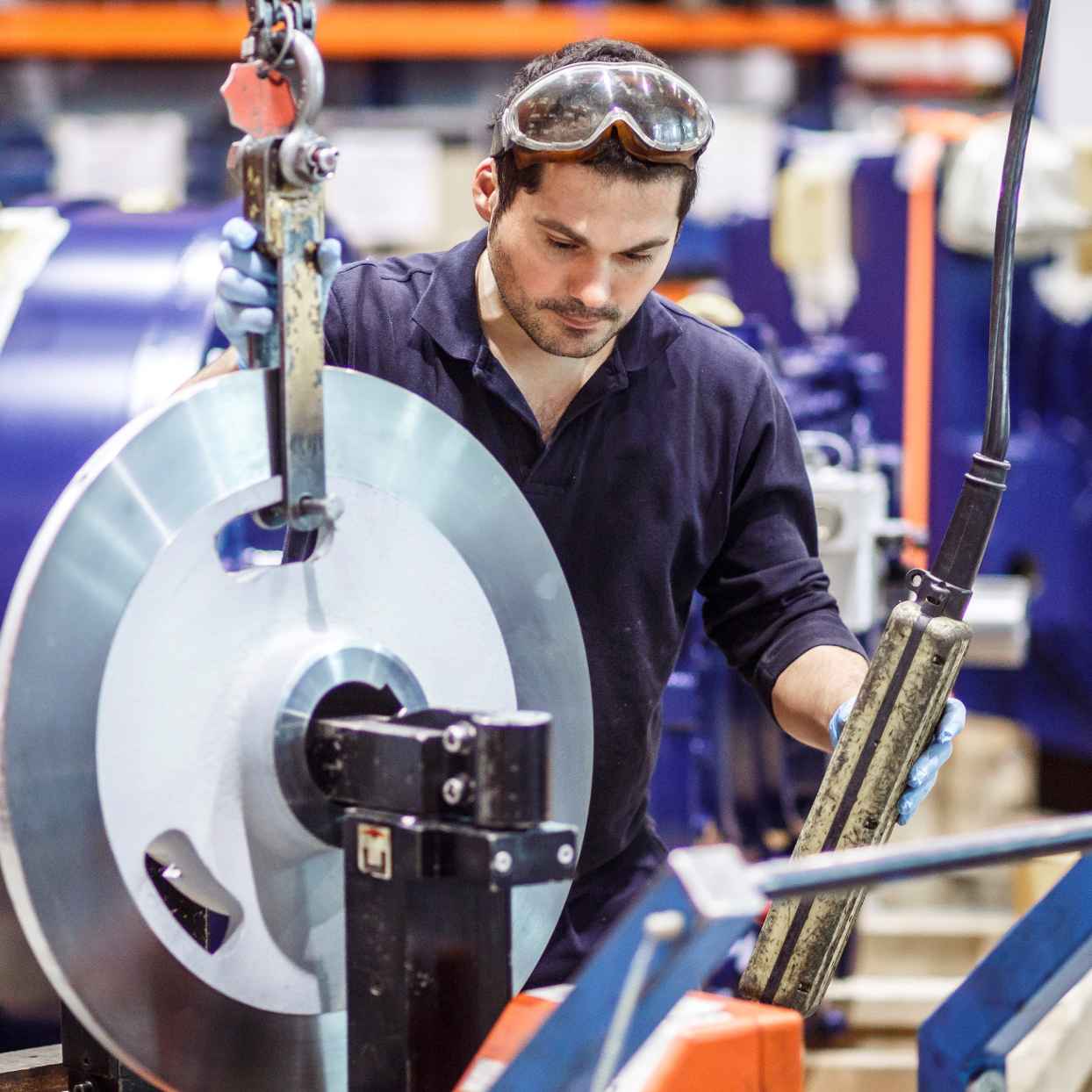Een medewerker met een veiligheidsbril verplaatst een groot metalen onderdeel in een fabriek. Op de achtergrond staan machines.