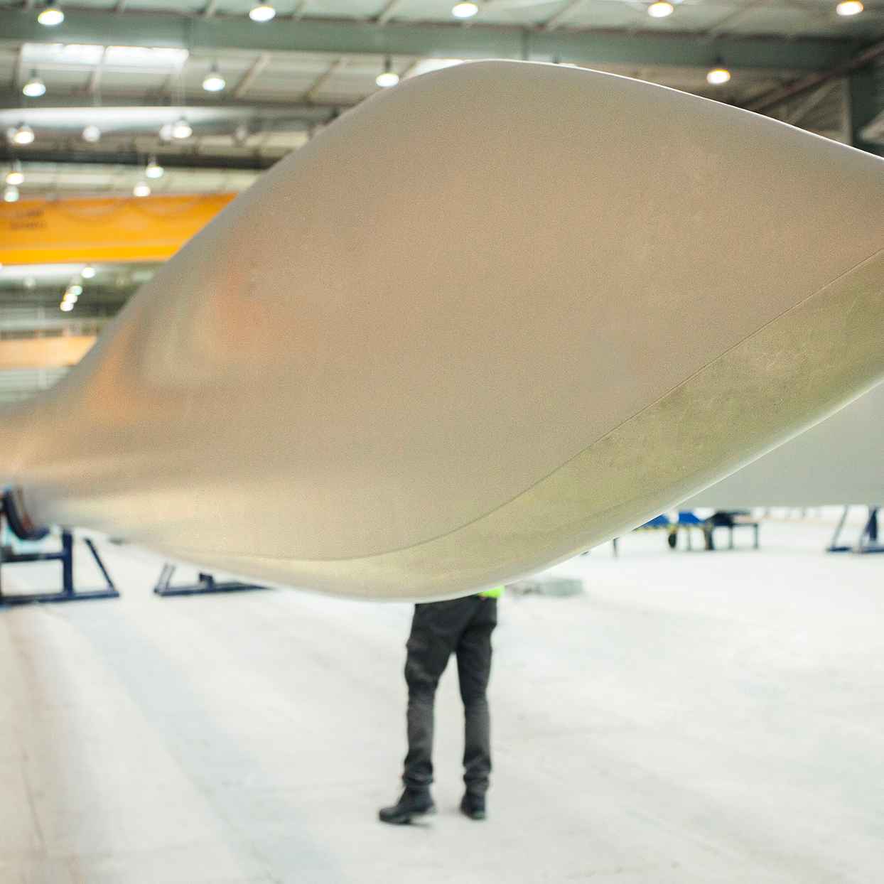 Een grote wiek van een windturbine ligt in een frabriekshal op steunen. Een persoon staat ernaast, waardoor de wiek extra groot lijkt.