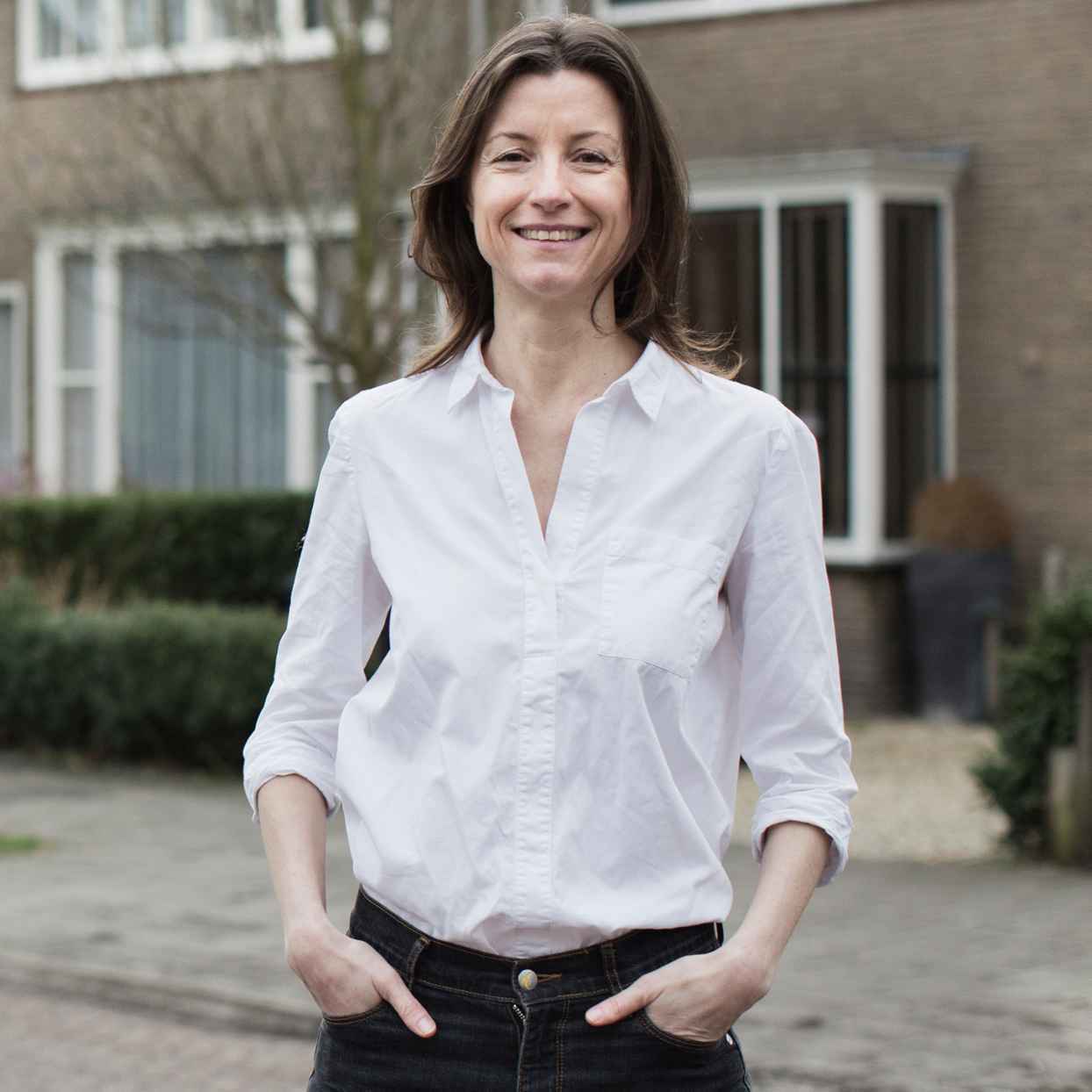 Een vrouw staat buiten in een woonwijk en draagt een witte blouse.