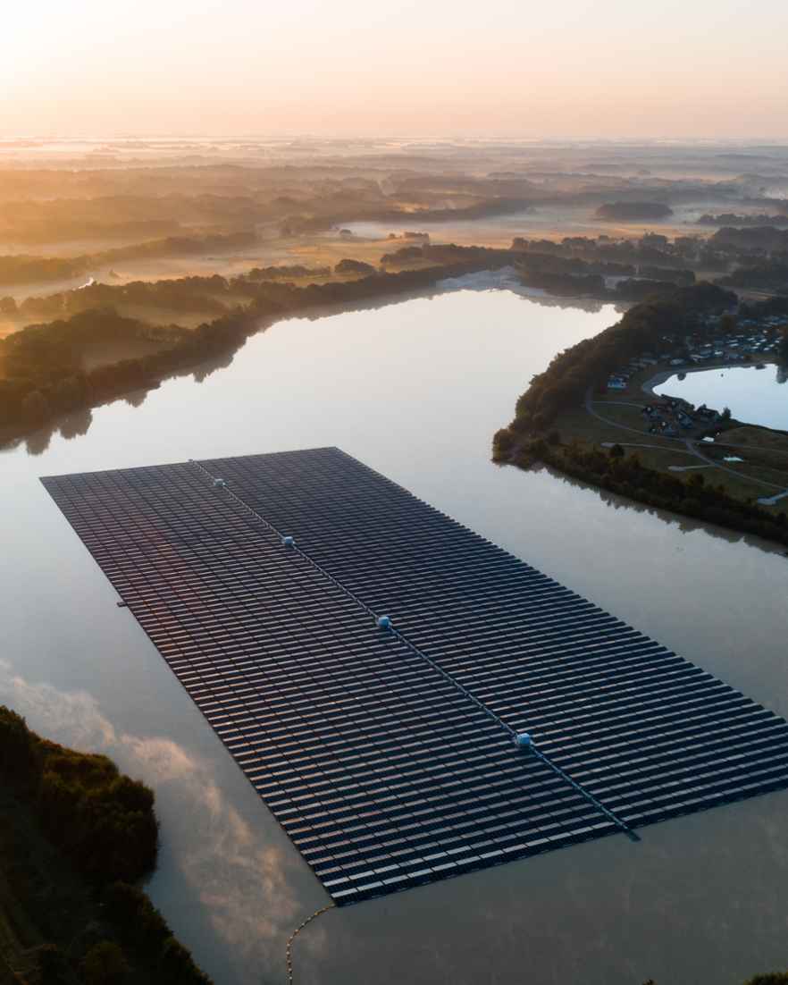 Luchtfoto van een groot veld met zonnepanelen naast water. In de verte liggen meer stroken land en water.